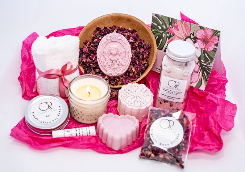 May include: A pink and white spa gift set with a candle, bath bombs, bath salts, rose petals, and a hand towel. The set is packaged in pink tissue paper and includes a card with a floral design.