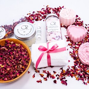 May include: A spa gift set with a pink and white theme. The set includes a soy candle, bath salts, a bath bomb, soap, a hand towel, and dried rose petals. The products are all packaged in clear glass jars and containers.