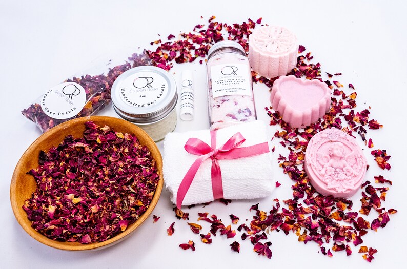 May include: A spa gift set with a pink and white theme. The set includes a soy candle, bath salts, a bath bomb, soap, a hand towel, and dried rose petals. The products are all packaged in clear glass jars and containers.