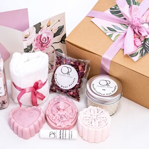 May include: A gift box filled with a range of bath and body products, including bath salts, a towel, soap, a candle, and rose petals. The box is wrapped in brown paper with a pink ribbon and a floral design.