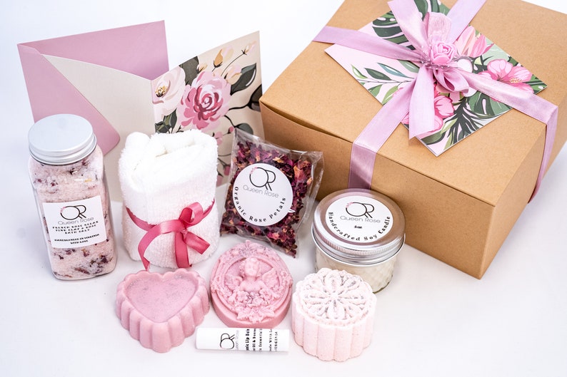 May include: A gift box filled with a range of bath and body products, including bath salts, a towel, soap, a candle, and rose petals. The box is wrapped in brown paper with a pink ribbon and a floral design.