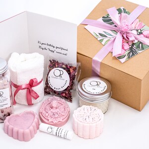 May include: A gift box filled with rose-themed bath and body products. The box is wrapped in brown paper with a pink ribbon and a floral design. The products include bath salts, a towel, rose petals, soap, and a lip balm. The box also includes a card with the text "It's your birthday, good friend. May the "happy" never end!"