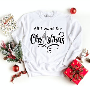 May include: White sweatshirt with the text "All I want for Christmas" in black cursive font. A hand with a middle finger extended is drawn in the middle of the text.
