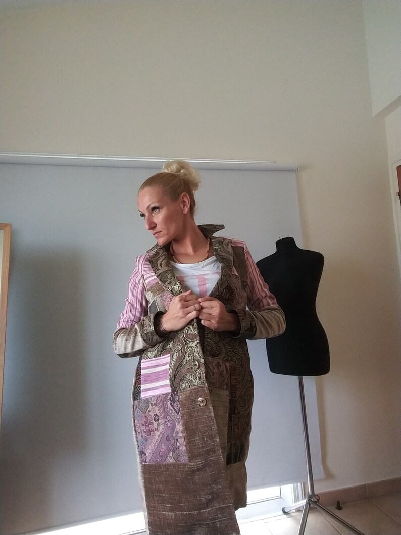 K&ouml;nnte beinhalten: Ein Patchwork-Mantel mit einer Mischung aus Mustern und Texturen. Der Mantel hat ein Paisley-Design, rosa gestreifte &Auml;rmel und einen braunen, strukturierten Stoff. Er hat einen Kragen und einen Knopfverschluss. Eine schwarze Schneiderpuppe steht im Hintergrund.