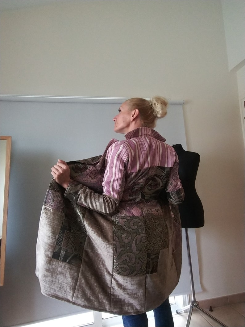 K&ouml;nnte beinhalten: Ein langer Patchwork-Mantel mit einer Mischung aus gemusterten und unifarbenen Stoffen in Braun-, Rosa- und Gr&uuml;nt&ouml;nen. Der Mantel hat einen gestreiften Kragen und &Auml;rmel mit einem einzigartigen Design. Der Mantel wird offen gehalten und zeigt das Innenfutter.