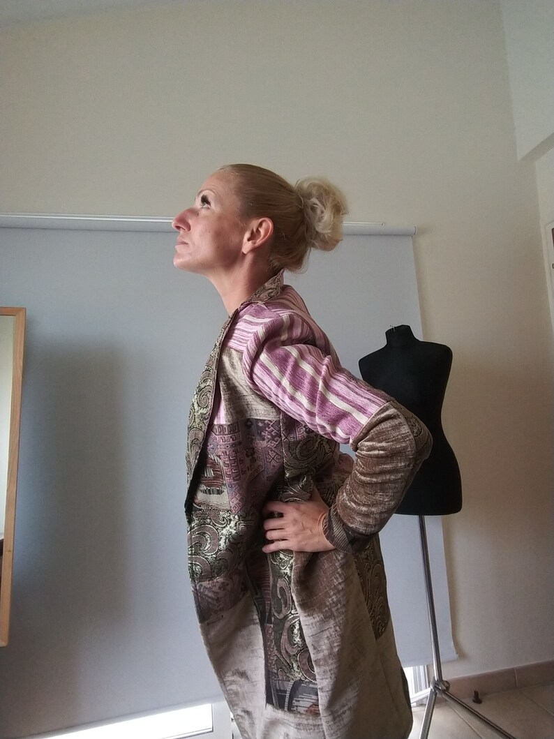 K&ouml;nnte beinhalten: Eine lange, gemusterte Jacke mit einer Mischung aus Farben, darunter Beige, Braun und lila Streifen. Die Jacke hat ein einzigartiges Design mit einer Mischung aus Texturen und Mustern. Die &Auml;rmel haben eine andere Farbe und ein anderes Muster als der K&ouml;rper der Jacke.