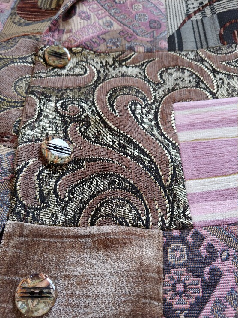 K&ouml;nnte beinhalten: Nahaufnahme eines Patchwork-Stoffs mit verschiedenen Mustern und Texturen. Das Design umfasst Paisley-, Blumen- und Streifenelemente in Braun-, Gold- und Rosat&ouml;nen. Dekorative Kn&ouml;pfe sind sichtbar.