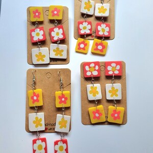 May include: Three pairs of dangle earrings with three square-shaped charms. Each charm features a white daisy with a yellow centre on a red, yellow, or white background. The earrings are handmade with love.