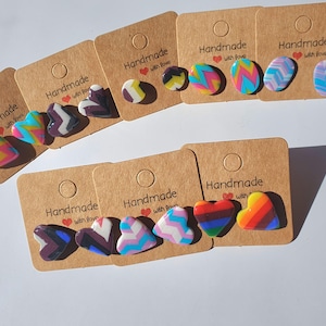 Puede incluir: Surtido de pendientes hechos a mano de arcilla polimérica en cartulina marrón con el texto "Handmade with love". Los pendientes tienen forma de corazón y óvalos con varios patrones coloridos.