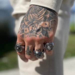 May include: Close-up of a hand adorned with four silver rings. The rings feature detailed designs, including a wolf, a lion, and other ornate patterns. The hand is heavily tattooed with a rose and the word "LOVE".