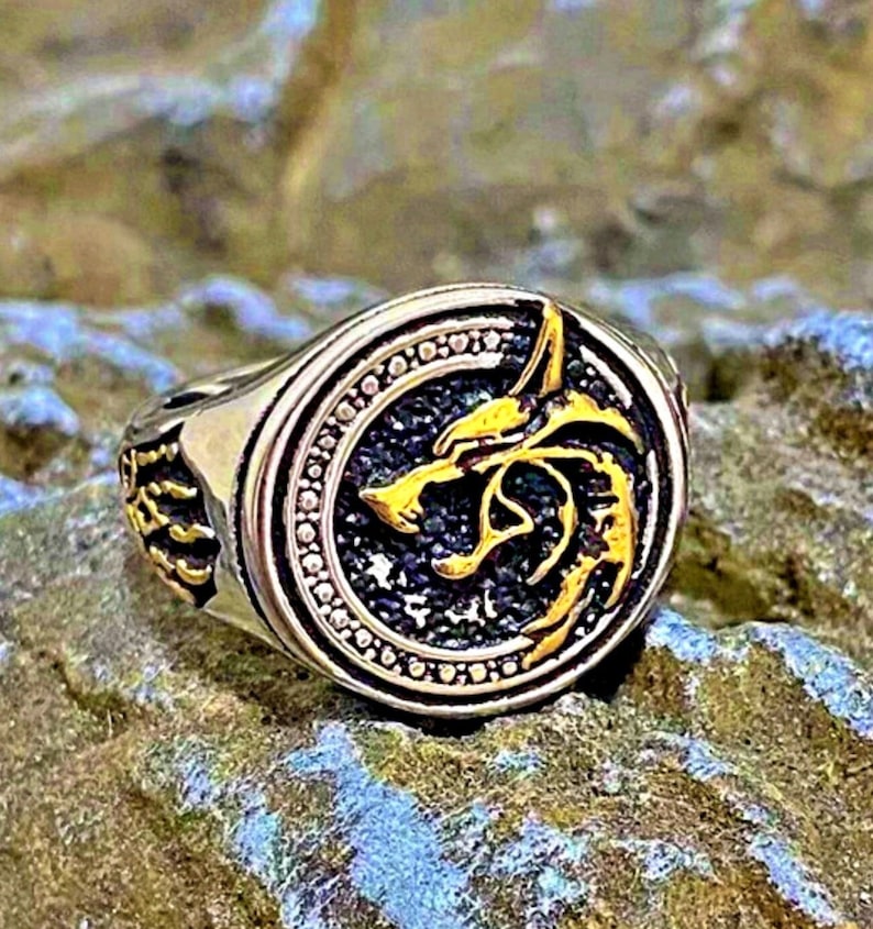 May include: A silver ring with a gold wolf head emblem. The ring has a detailed design with a black background and a circular border. The wolf head is in gold, and the ring is set on a stone surface.