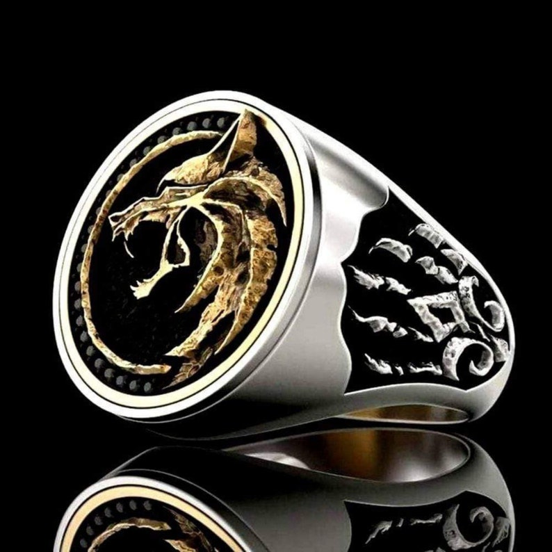 May include: A silver ring with a gold wolf head emblem on a black background. The ring has a detailed design with a textured wolf and decorative accents. The ring is a bold statement piece.