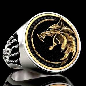 May include: A silver ring with a gold wolf head design on a black background. The ring has a gold border and a detailed wolf head. The band of the ring has a decorative design.