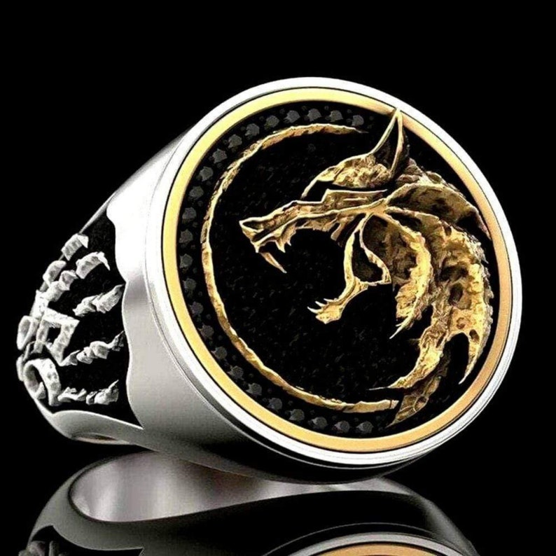 May include: A silver ring with a gold wolf head design on a black background. The ring has a gold border and a detailed wolf head. The band of the ring has a decorative design.
