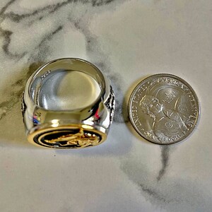 May include: A silver and gold-toned ring with an oval-shaped design. The ring is next to a quarter coin for size comparison. The ring has a detailed design on the sides and a gold-colored interior.
