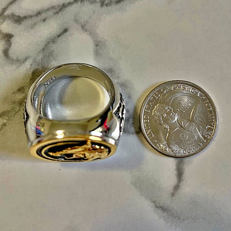 May include: A silver and gold-toned ring with an oval-shaped design. The ring is next to a quarter coin for size comparison. The ring has a detailed design on the sides and a gold-colored interior.