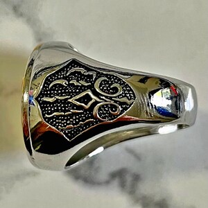 May include: A silver-toned ring with an ornate, black, shield-shaped design. The ring's band is smooth and polished, reflecting light. The shield features a textured pattern with swirling and geometric elements, creating a detailed and eye-catching accessory.