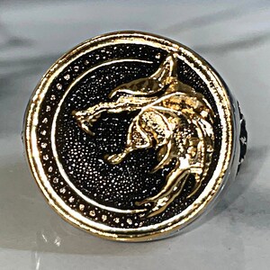 May include: A silver ring with a gold-colored wolf head design. The wolf is centered on a black background, surrounded by a gold-colored border. The ring has a detailed, textured appearance.