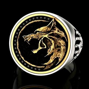 May include: A silver ring with a gold wolf head design on a black background. The wolf head is detailed and has an open mouth. The ring has a gold border and a textured silver band.