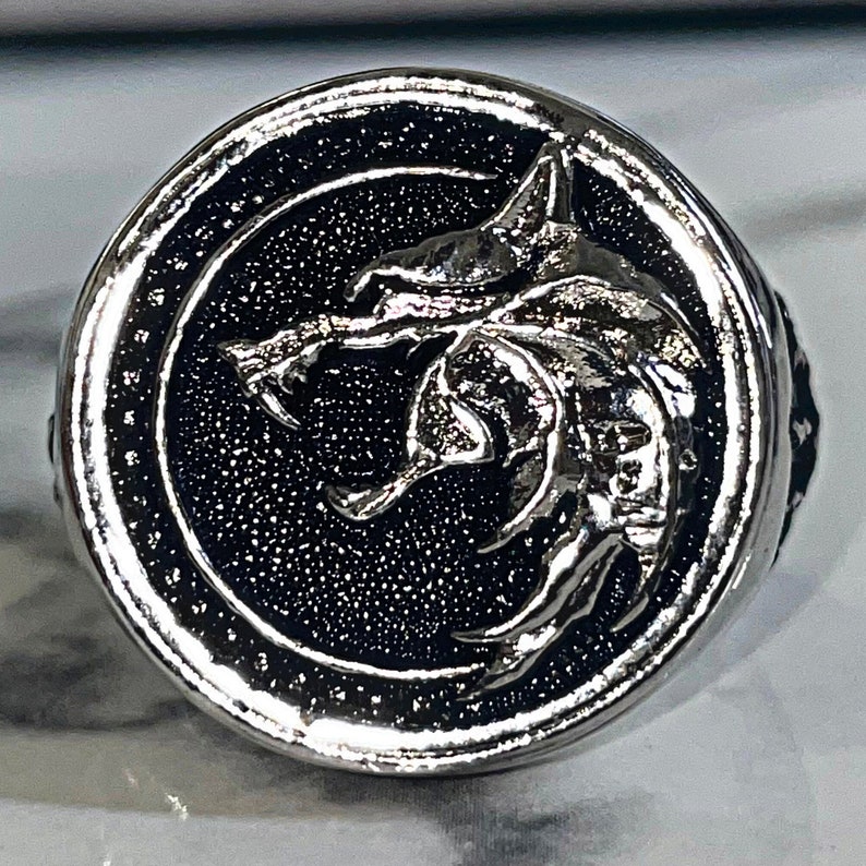 May include: Silver-toned ring featuring a detailed wolf head emblem set against a textured black background. The ring has a circular design with a raised wolf head in the center, and a polished silver border.