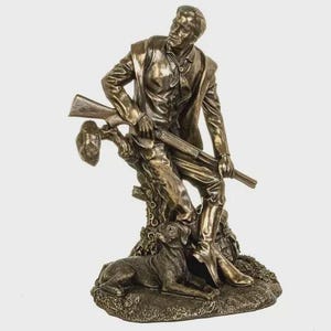 Può includere: Statua in bronzo di un uomo in giacca e stivali, con un fucile, seduto su un tronco d'albero. Un cane è sdraiato alla base dell'albero. La statua ha una finitura antica e dettagliata.