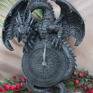 May include: A dark gray dragon-shaped clock with Roman numerals. The dragon has detailed wings and scales, perched atop a clock face with a Celtic knot design. The clock hands point to approximately 6 o'clock. Decorative holiday accents are visible.