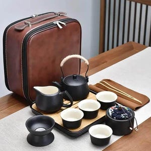 Puede incluir: Un juego de té portátil que incluye una tetera de cerámica negra mate con asa envuelta en cuerda, una jarra a juego, cuatro tazas pequeñas, un colador y un recipiente con tapa, sobre una bandeja de madera dentro de un estuche marrón.
