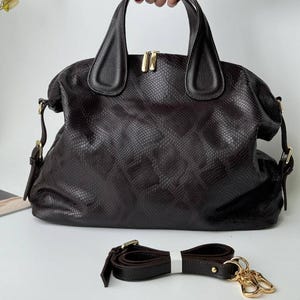 Peut inclure: Un sac à main marron foncé avec un motif texturé en peau de serpent, doté de deux poignées supérieures, d'une fermeture éclair double dorée et d'attaches latérales. Une bandoulière amovible assortie avec des mousquetons dorés est incluse.