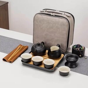 Puede incluir: Un juego de té portátil que incluye una tetera negra mate, una jarra a juego y cuatro tazas pequeñas con interior blanco. El set contiene una bandeja de madera, pinzas de bambú, un recipiente negro con tapa floral y un estuche gris.
