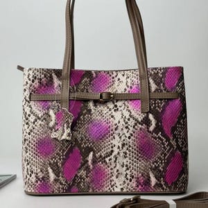 Op de afbeelding: Een tote bag met een levendig slangenleerpatroon in paars en beige. De tas heeft twee bruine schouderbanden, een horizontale bruine riem met een goudkleurige gesp aan de voorzijde en een bijpassende afneembare bruine schouderband.