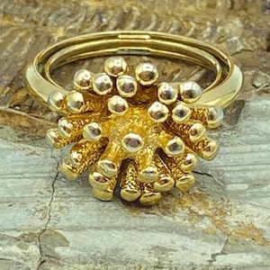 May include: A gold-colored ring with a unique design. The ring features a double band and a central cluster of small, round, gold-colored beads, creating a textured, floral-like appearance. The ring is set against a textured, gray and brown background.
