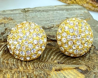 May include: A pair of gold-toned, round stud earrings encrusted with numerous clear crystals. The earrings are set against a textured, gray-brown stone surface, creating a contrast between the sparkle of the jewelry and the natural backdrop.