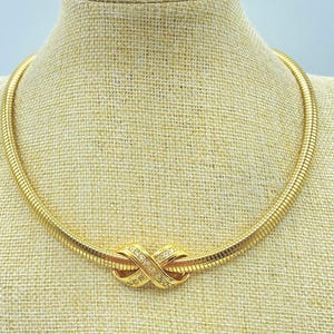 May include: A gold-toned necklace with a textured, ribbed design. The necklace features a knot-shaped centerpiece adorned with small, clear gemstones. The jewelry is displayed on a beige mannequin, showcasing its design and style.