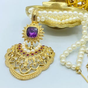 Pode incluir: Um colar de tom dourado com um fio de p&eacute;rolas brancas artificiais e um pendente grande e ornamentado. O pendente apresenta uma pedra quadrada roxa central, rodeada por pequenos cristais vermelhos e transparentes numa arma&ccedil;&atilde;o de metal dourado.