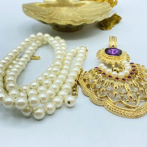 Pode incluir: Um colar de tom dourado com um fio de p&eacute;rolas brancas artificiais e um pendente grande e ornamentado. O pendente &eacute; decorado com uma pedra central roxa, pequenas p&eacute;rolas brancas e cristais vermelhos e transparentes em metal filigranado.
