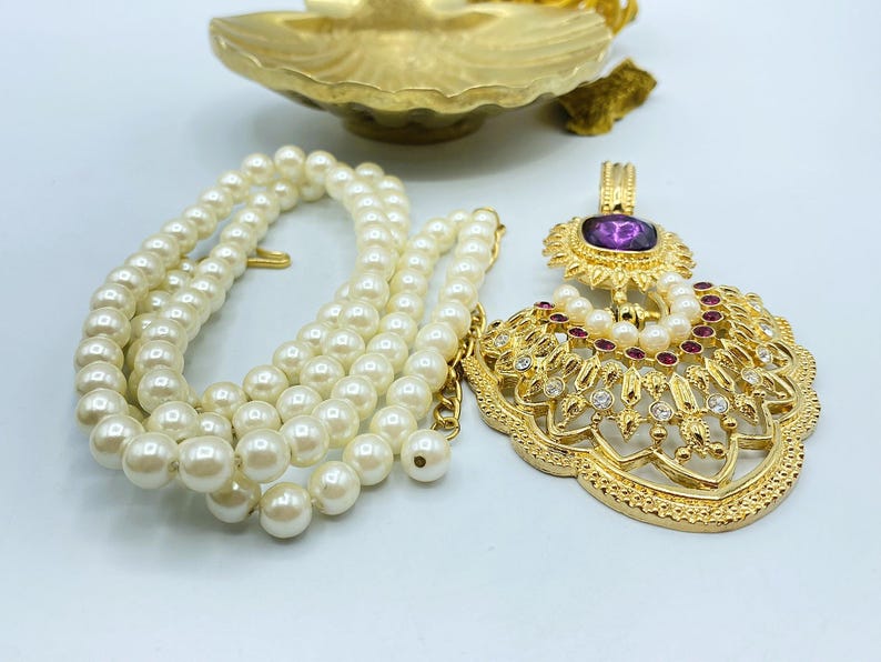 Pode incluir: Um colar de tom dourado com um fio de p&eacute;rolas brancas artificiais e um pendente grande e ornamentado. O pendente &eacute; decorado com uma pedra central roxa, pequenas p&eacute;rolas brancas e cristais vermelhos e transparentes em metal filigranado.