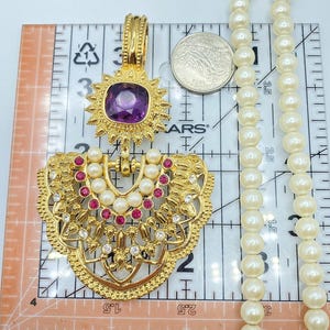 Pode incluir: Um pendente dourado com uma pedra preciosa roxa central, pequenas p&eacute;rolas brancas e strass rosa e transparentes, preso a um longo colar de p&eacute;rolas brancas. Uma moeda e uma r&eacute;gua mostram uma altura de 7,6 cm.