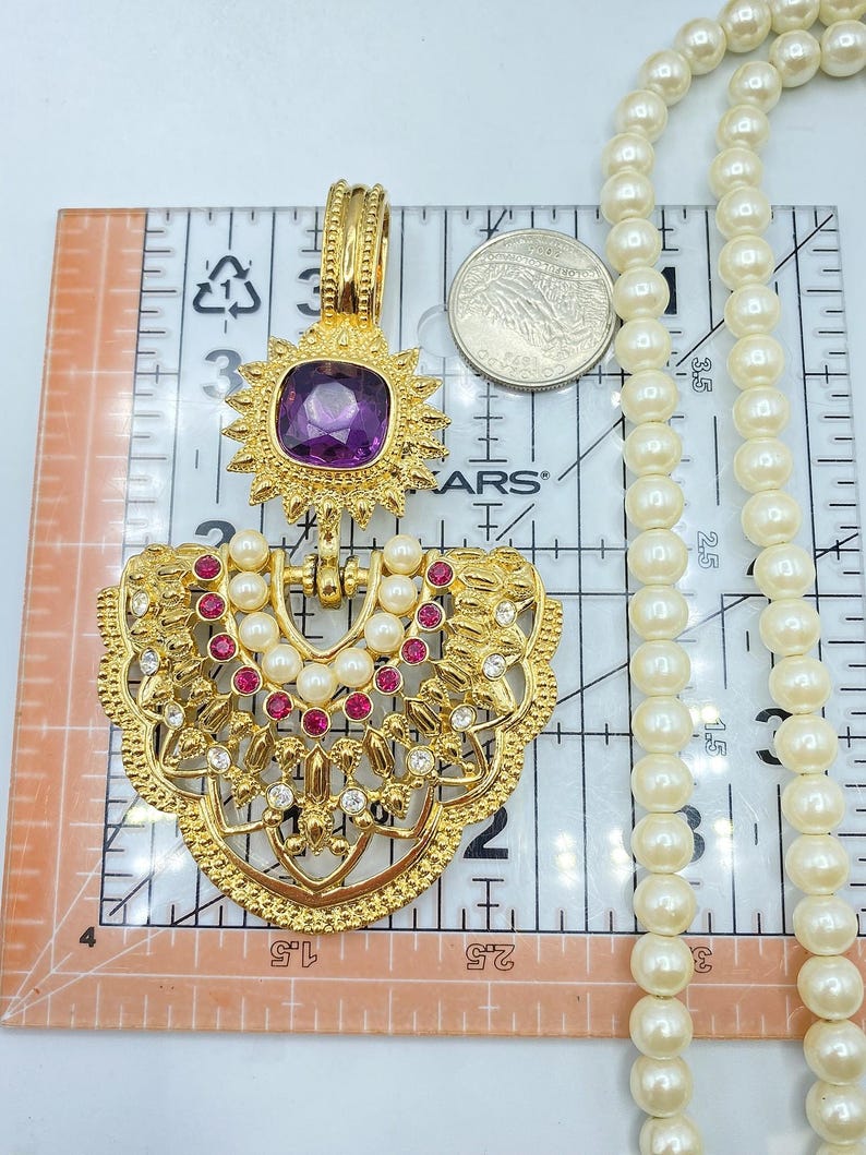 Pode incluir: Um pendente dourado com uma pedra preciosa roxa central, pequenas p&eacute;rolas brancas e strass rosa e transparentes, preso a um longo colar de p&eacute;rolas brancas. Uma moeda e uma r&eacute;gua mostram uma altura de 7,6 cm.