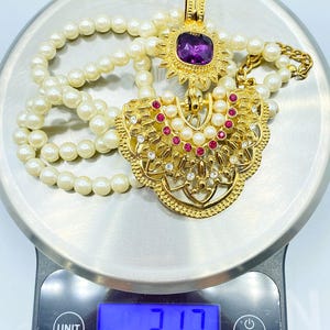 Pode incluir: Um colar com pendente em tom dourado com uma grande pedra preciosa quadrada roxa, pequenos strass vermelhos e transparentes e p&eacute;rolas artificiais brancas. O colar repousa sobre uma balan&ccedil;a digital que indica um peso de 89,87 gramas.