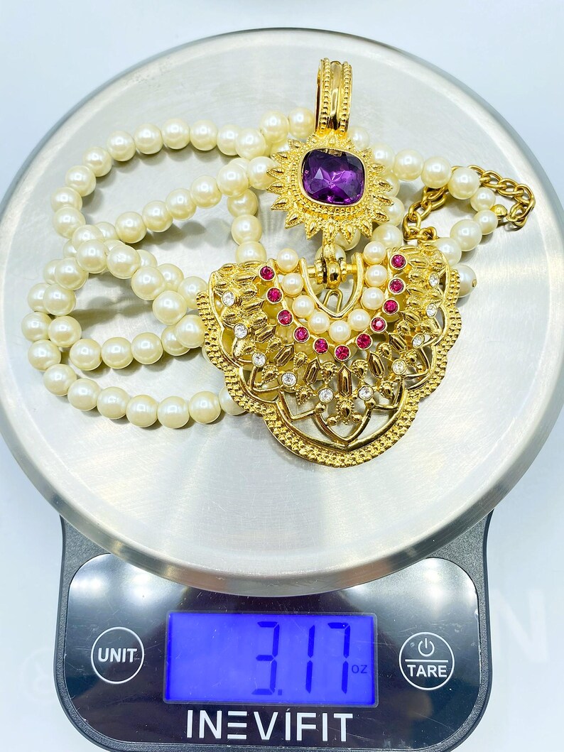 Pode incluir: Um colar com pendente em tom dourado com uma grande pedra preciosa quadrada roxa, pequenos strass vermelhos e transparentes e p&eacute;rolas artificiais brancas. O colar repousa sobre uma balan&ccedil;a digital que indica um peso de 89,87 gramas.