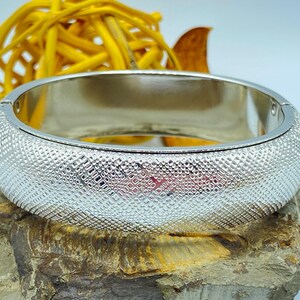 May include: A silver-colored bangle bracelet with a textured, diamond-patterned surface. The bracelet is hinged and appears to be made of metal. The background includes a yellow decorative element and a stone.