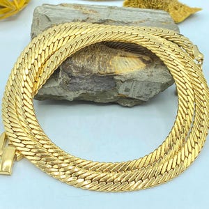 May include: A gold-toned herringbone chain necklace featuring a flat, woven link design with a polished finish. The necklace is arranged in a circular coil and includes a rectangular clasp mechanism at one end, showcasing a sleek, metallic appearance.