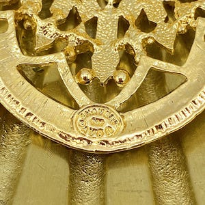Pode incluir: Um broche de metal em tom dourado com um design intrincado de filigrana e acabamento texturizado. A pe&ccedil;a inclui um selo circular na base com o texto AVON claramente vis&iacute;vel, indicando o fabricante deste item de acess&oacute;rio decorativo.