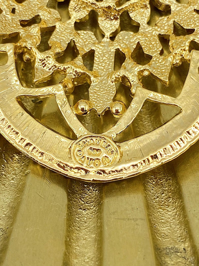 Pode incluir: Um broche de metal em tom dourado com um design intrincado de filigrana e acabamento texturizado. A pe&ccedil;a inclui um selo circular na base com o texto AVON claramente vis&iacute;vel, indicando o fabricante deste item de acess&oacute;rio decorativo.