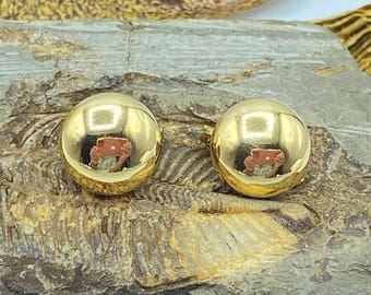 May include: Two round, gold-colored earrings. Each earring has a smooth, reflective surface, with a mirrored reflection of the surroundings. The earrings are set against a textured, natural background.
