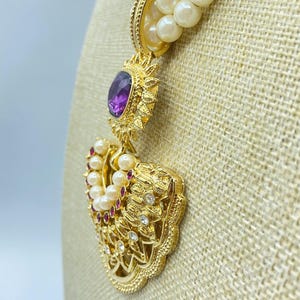 Pode incluir: Um colar de p&eacute;rolas de v&aacute;rias voltas com um pendente dourado que apresenta uma pedra central oval roxa. O pendente &eacute; decorado com pequenas p&eacute;rolas brancas, pedras vermelhas e cristais transparentes numa arma&ccedil;&atilde;o de metal dourado texturado.