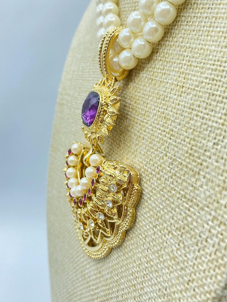 Pode incluir: Um colar de p&eacute;rolas de v&aacute;rias voltas com um pendente dourado que apresenta uma pedra central oval roxa. O pendente &eacute; decorado com pequenas p&eacute;rolas brancas, pedras vermelhas e cristais transparentes numa arma&ccedil;&atilde;o de metal dourado texturado.
