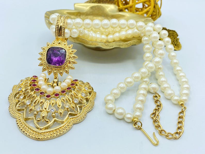 Pode incluir: Um colar de tom dourado com um fio de p&eacute;rolas brancas artificiais e um pendente grande e ornamentado. O pendente apresenta uma pedra quadrada roxa central, rodeada por pequenos cristais vermelhos e transparentes numa arma&ccedil;&atilde;o de metal dourado.