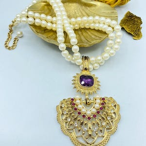 Pode incluir: Um colar de p&eacute;rolas brancas de duas voltas com um pendente de metal dourado. O pendente inclui uma pedra quadrada roxa central, uma moldura em forma de sol e uma base em leque adornada com pequenas p&eacute;rolas, pedras cor-de-rosa e cristais.