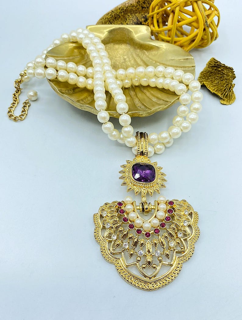 Pode incluir: Um colar de p&eacute;rolas brancas de duas voltas com um pendente de metal dourado. O pendente inclui uma pedra quadrada roxa central, uma moldura em forma de sol e uma base em leque adornada com pequenas p&eacute;rolas, pedras cor-de-rosa e cristais.